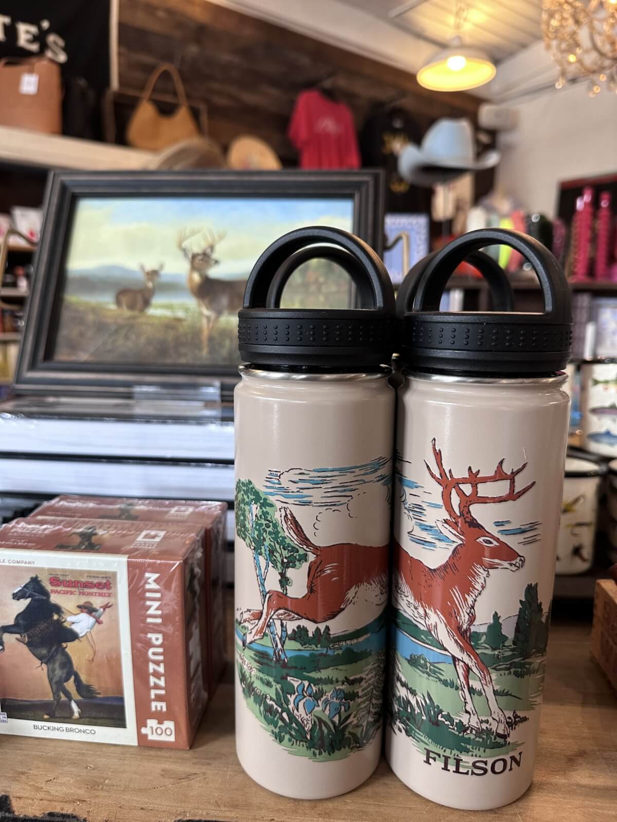 Two Filson water bottles with deer illustrations are displayed on a wooden counter alongside a puzzle box and a framed painting of deer in the background—perfect June FINDS for any Nashville adventurer.