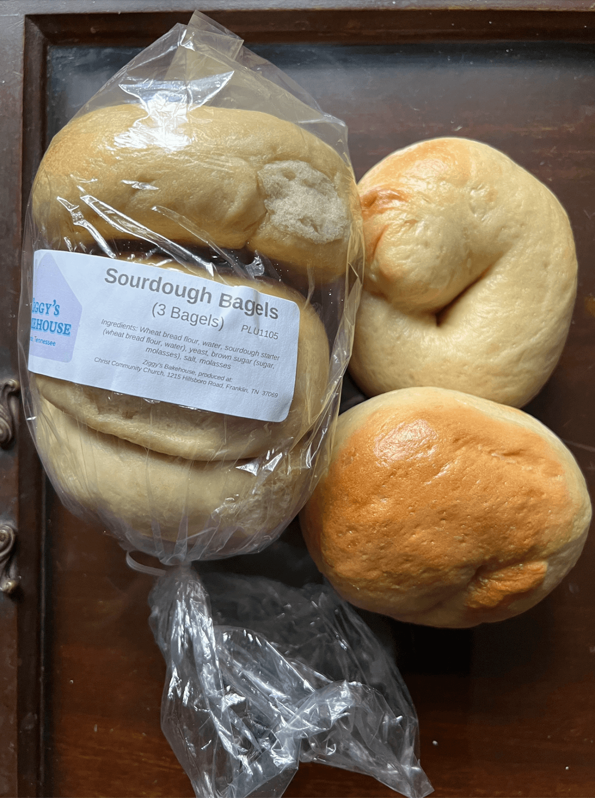 Three plain sourdough bagels, two loose and one in a partially opened plastic package labeled “Sourdough Bagels (3 Bagels),” rest on a wooden surface—an inviting Nashville FINDS pick for June.