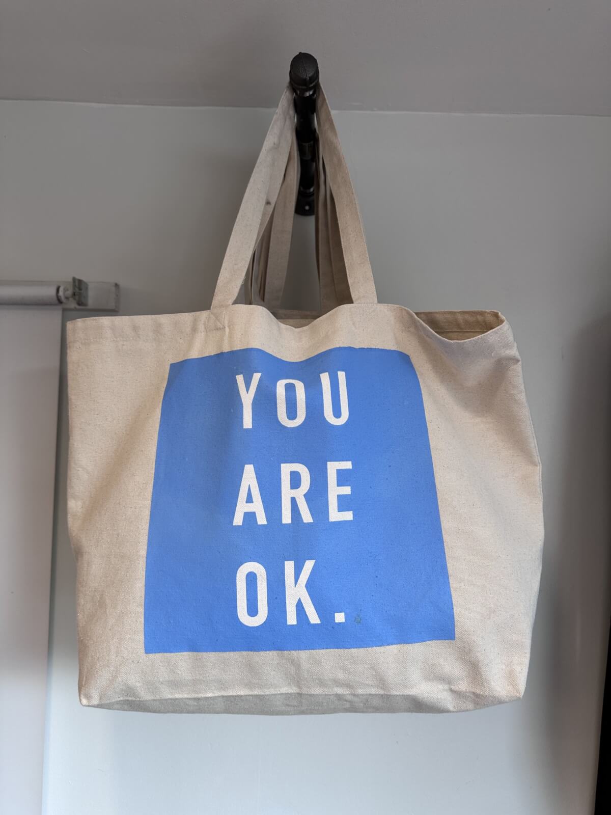 A beige canvas tote bag from our June FINDS collection, featuring a blue rectangle and the words "YOU ARE OK." printed in white capital letters—a stylish nod to Nashville cool.