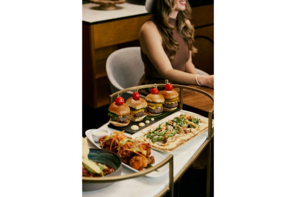 A woman sits at a table at Mason Bar with a serving cart of July 4th specials, featuring mini burgers with cherry tomatoes, flatbread pizza, and a dish of fried chicken with salad.