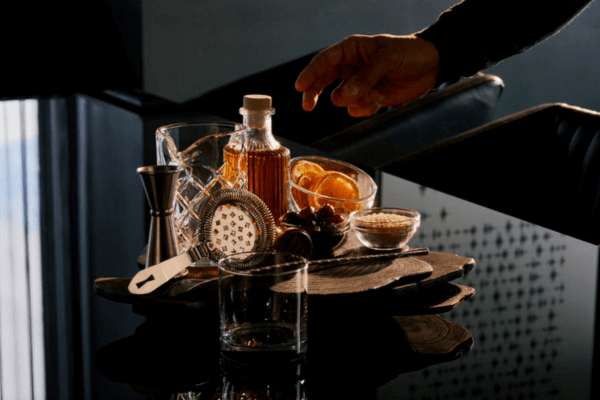 A hand reaches over a tray holding a glass, a bottle of bourbon, a strainer, bar tools, sliced oranges, grapes, and a bowl of sugar on a dark table—perfect for crafting cocktails in style, no bowties required.