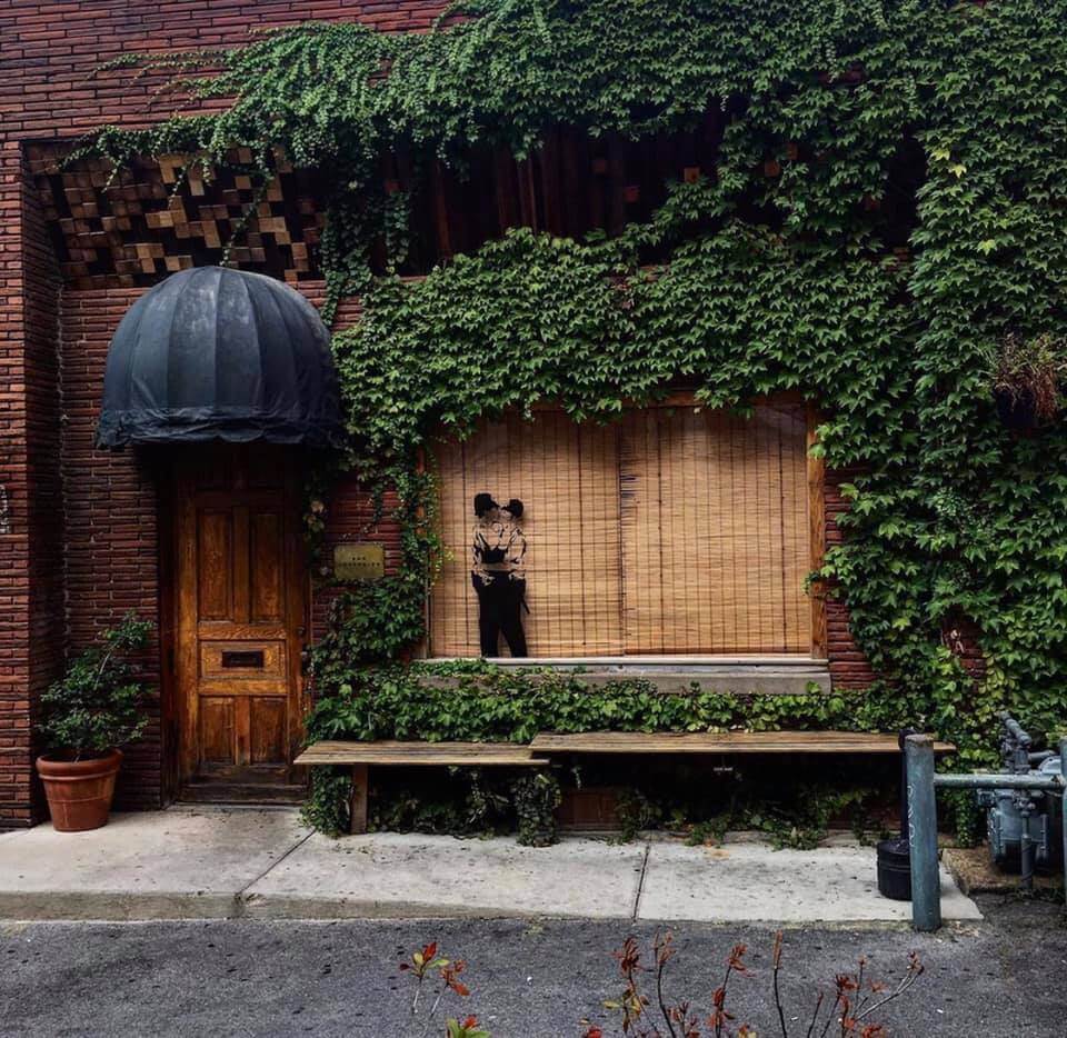A brick building covered in ivy with a wooden door, a black awning, a bench, and a mural of two people embracing on the window—a charming Nashville spot perfect for music and meal pairings.