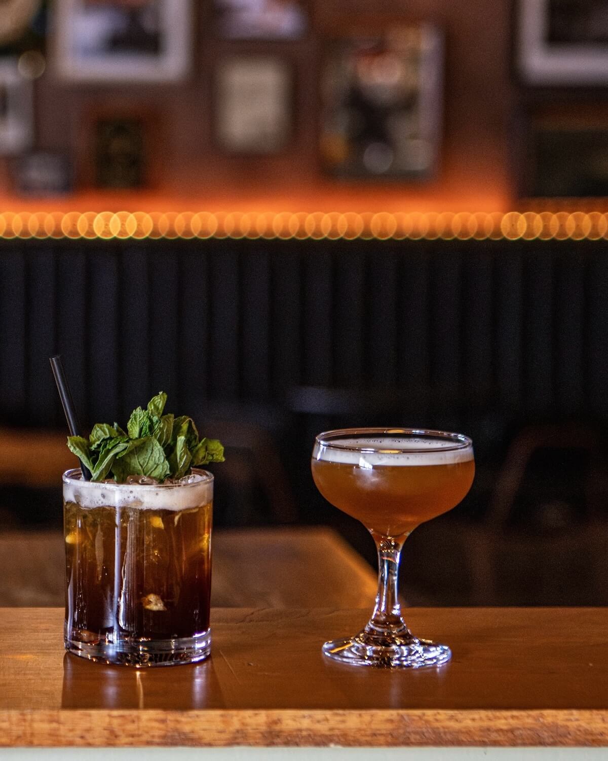 Two cocktails on a wooden bar in Nashville: one in a short glass with mint garnish and a straw, the other in a coupe glass with light foam. Blurred background evokes warm lighting, perfect for conversations about local music venues and restaurants.