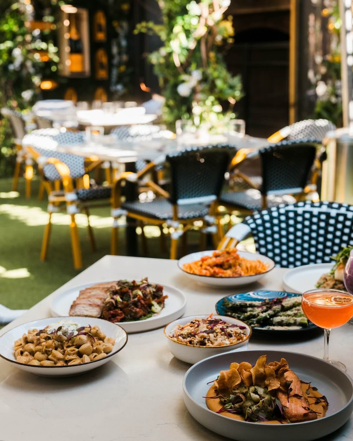 A table set with various dishes and a cocktail in an outdoor restaurant, surrounded by green plants and empty chairs—perfect for those looking to eat near Nashville music venues.