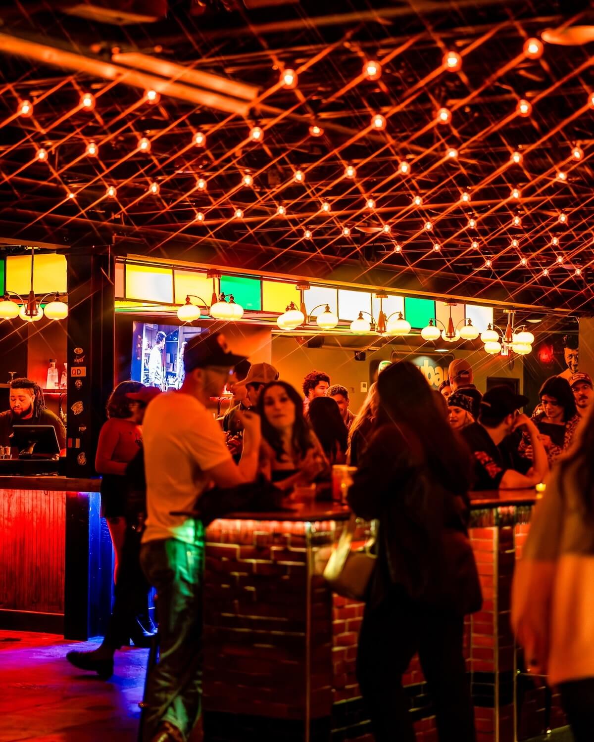 A crowded Nashville bar with colorful lights and people socializing around the counter under warm, orange string lights, enjoying lively music pairings.