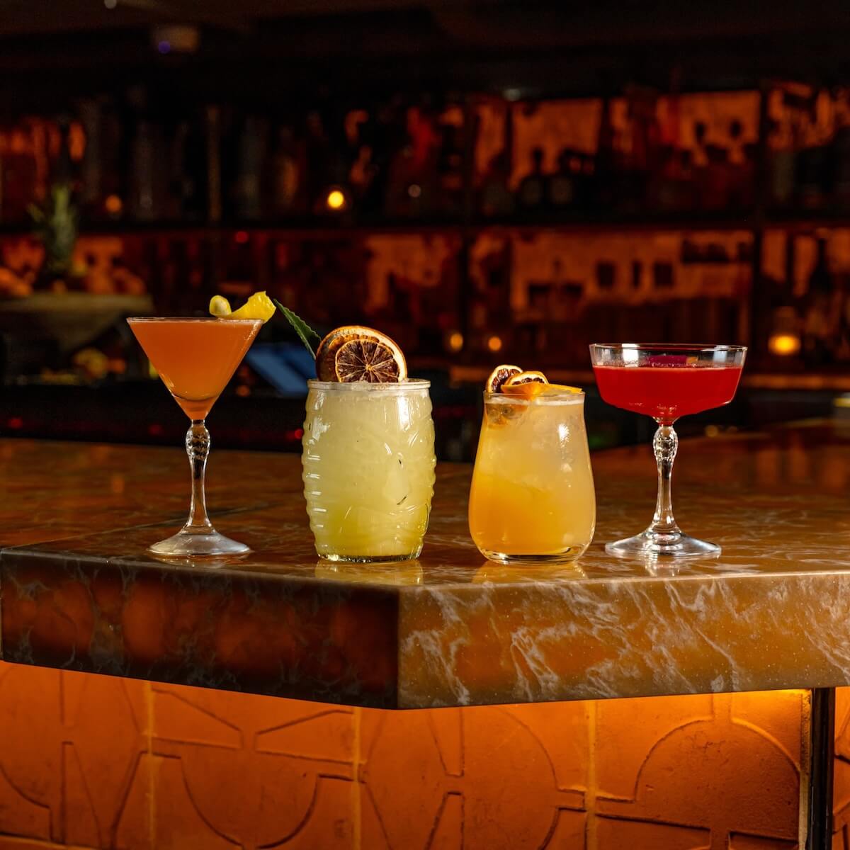 Four colorful cocktails sit on a marble bar counter in a dimly lit Nashville setting, each garnished with fruit.