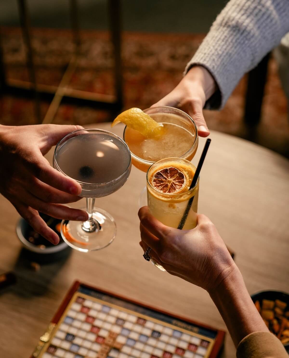 Three hands toast colorful cocktails over a Scrabble board, capturing the lively spirit found in Nashville music venues.