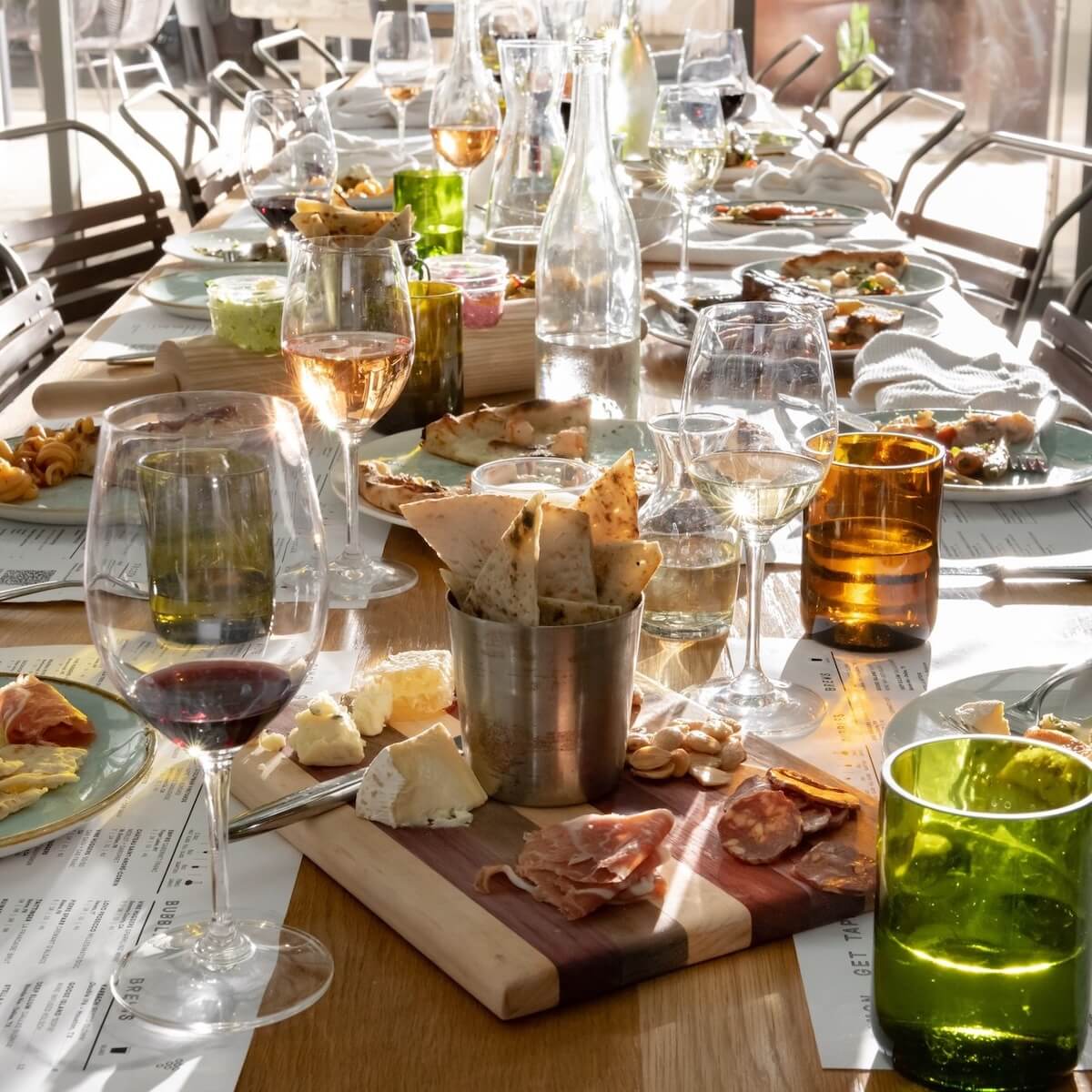 A sunlit dining table in Nashville set with wine glasses, water bottles, assorted cheeses, charcuterie, bread, and various appetizers awaits guests for a feast of meal pairings and lively conversation.
