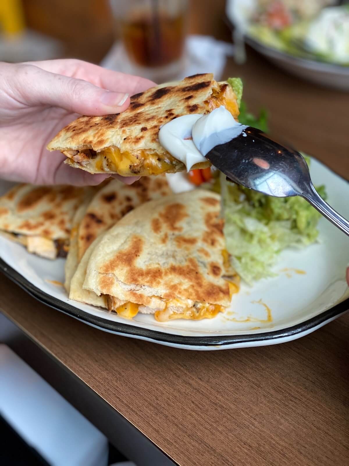 A hand holds a cheesy quesadilla above a plate as a spoon adds a dollop of sour cream; just like the bites you might enjoy before catching live acts at Nashville music venues, with more quesadilla slices and guacamole on the plate.