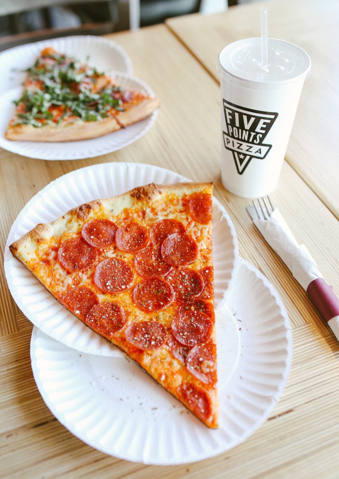 A slice of pepperoni pizza on a paper plate, a cup with a straw labeled "Five Points Pizza," a napkin, and another pizza slice in the background on a wooden table—classic Nashville meal pairings at their best.