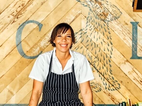 Chef Nina Compton, in a striped apron and white shirt, stands smiling before a wooden wall adorned with a large rabbit illustration and partially visible letters.