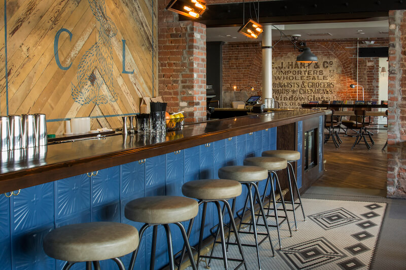 A modern bar with a blue tile front, metal stools, wooden countertop, exposed brick walls, and vintage signs in an industrial-style interior—perfect for savoring cocktails.