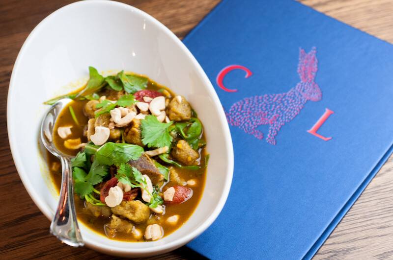 A bowl of curry garnished with cilantro and peanuts sits next to a blue menu with a red rabbit illustration and the letters C and L, reflecting the vibrant style of chef Nina Compton.