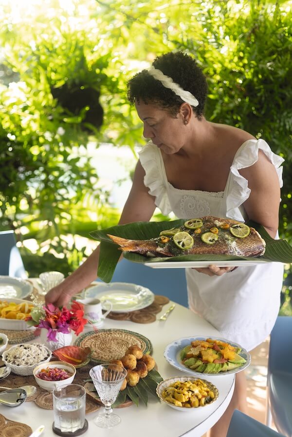 Chef Nina Compton, in a white dress, places a platter of whole fish with lemon slices on a table set with various dishes outdoors, surrounded by lush greenery.