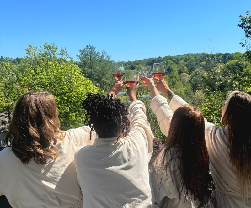 Four people in robes raise glasses of wine together on a balcony overlooking a green, tree-covered landscape under a clear blue sky.