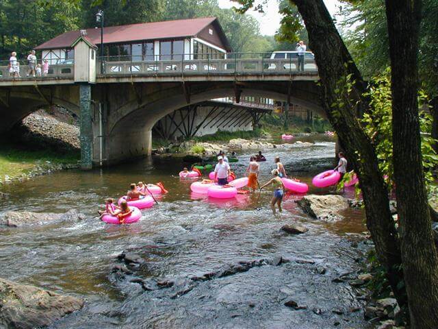 People float and wade in a shallow river with pink inner tubes near a bridge and a building.