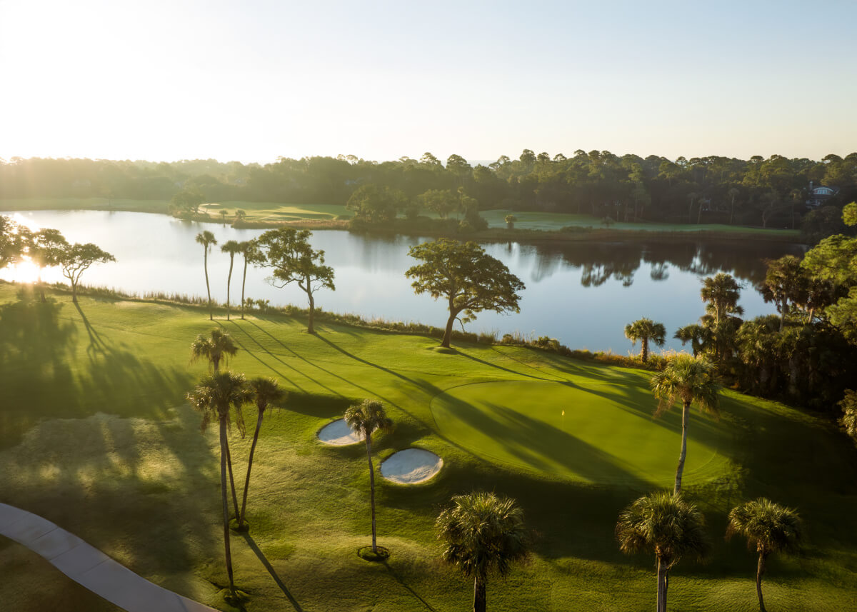 A picturesque golf course with sand bunkers, palm trees, and a lake, photographed at sunrise with long shadows—perfect for summer weekend trips.