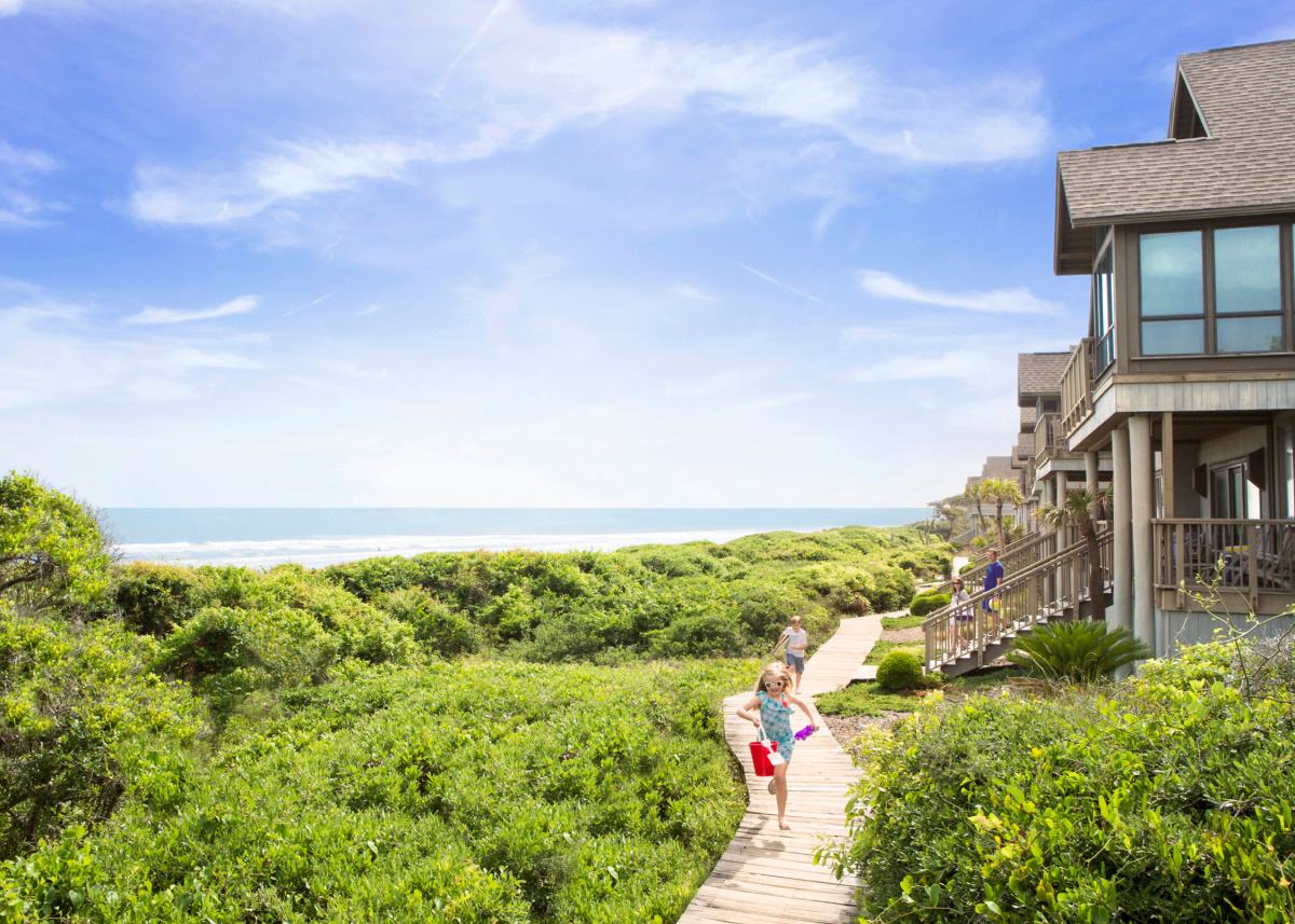 Children walk along a wooden boardwalk through green dunes toward the beach, perfect for summer weekend trips, with seaside houses and a blue sky in the background.