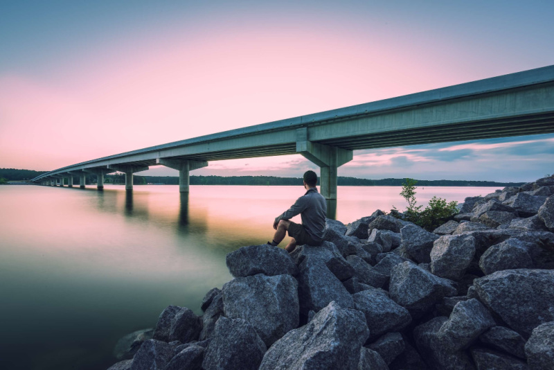 A person sits on rocks by the water, gazing at a long bridge stretching across a calm river under a pastel sunset sky—an ideal scene for those seeking peaceful weekend trips from Nashville.