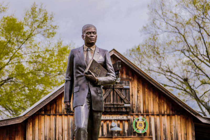 A bronze statue of a man in a suit holding a book stands before a rustic wooden building with trees and a cloudy sky—an intriguing stop for those seeking unique weekend trips from Nashville.