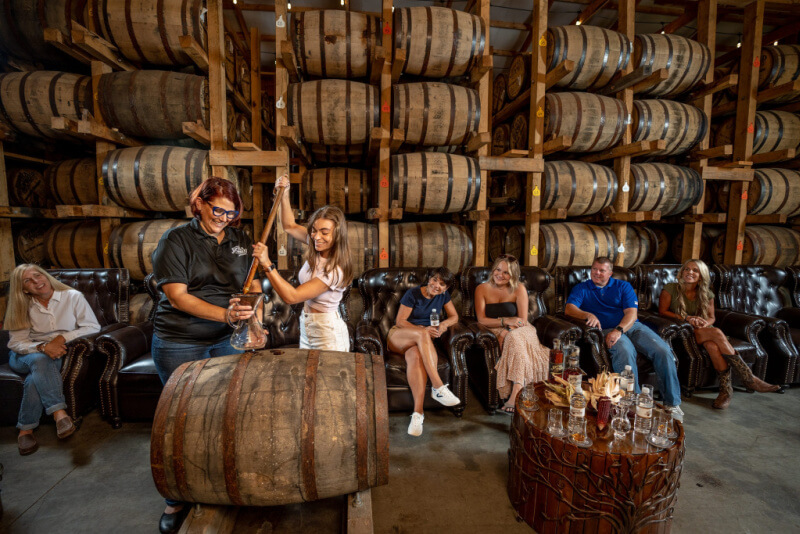 Two women pour whiskey from a barrel as several people watch, seated on leather sofas in a warehouse filled with stacked wooden barrels—a perfect stop on weekend trips from Nashville.