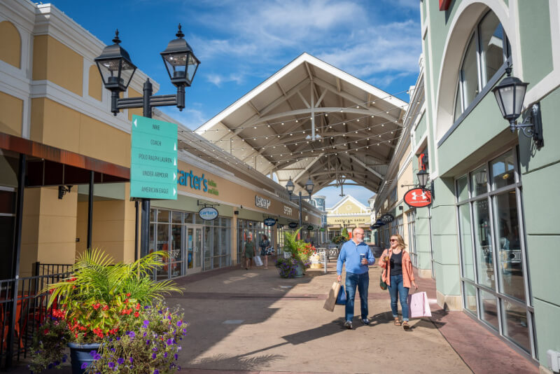 Two people stroll through an outdoor shopping center with stores on both sides, carrying shopping bags under a partially covered walkway on a sunny day—perfect for weekend trips from Nashville.