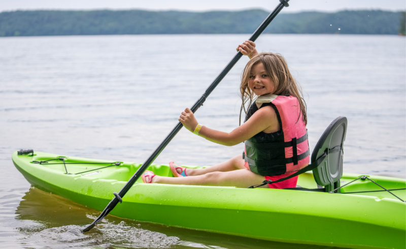 A young girl wearing a life jacket paddles a green kayak on a calm lake near Nashville, with forested hills in the background—a perfect scene for summer weekend trips.