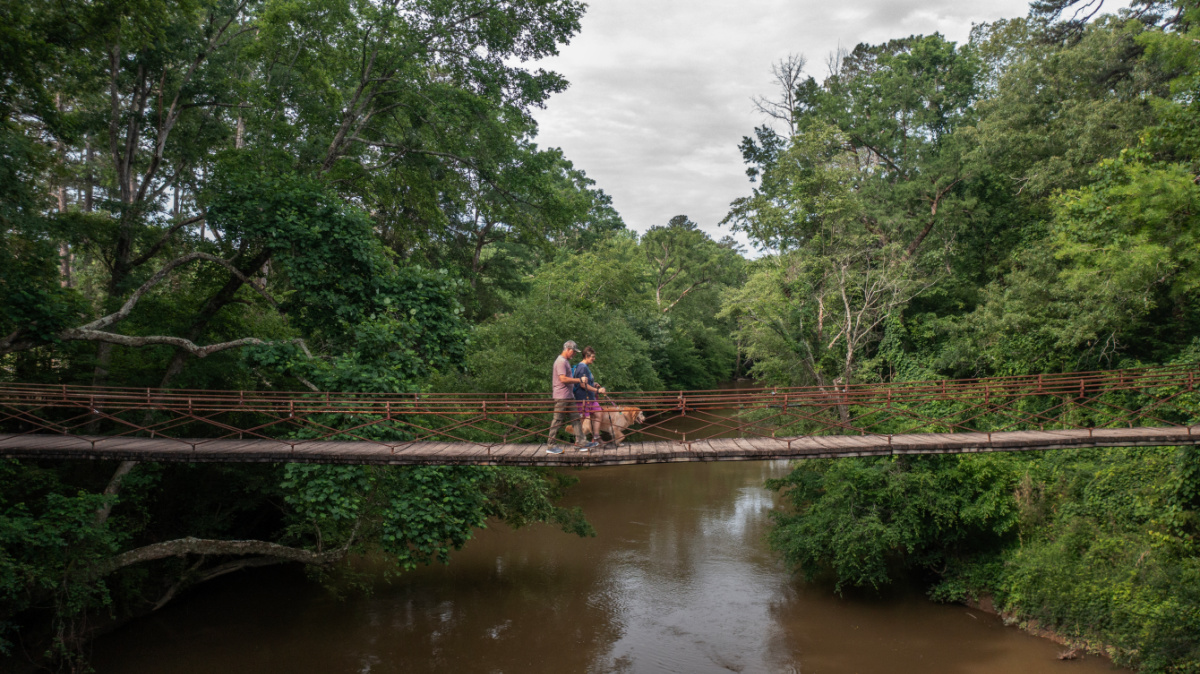 Three people stand on a narrow suspension bridge over a brown river, surrounded by dense green trees and cloudy sky—a perfect snapshot from summer weekend trips near Nashville.
