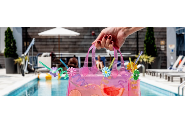 A hand with painted nails holds a translucent pink bag decorated with colorful straws and umbrellas by a swimming pool, ready for the ultimate Pool Party.