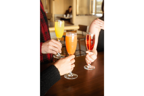 Three people clink glasses filled with orange, yellow, and red beverages while sitting at a wooden table in a well-lit room at The Restaurant during Drag Brunch.
