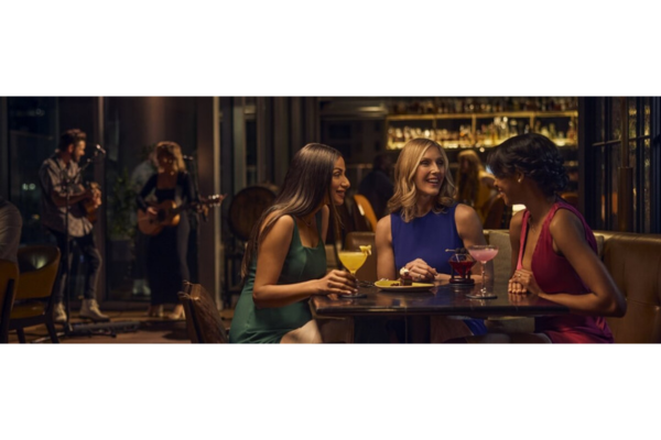 Three women sit at a table with drinks, talking and smiling in the dimly lit Thistle and Rye bar while a band performs during Writers’ Rounds in the background.