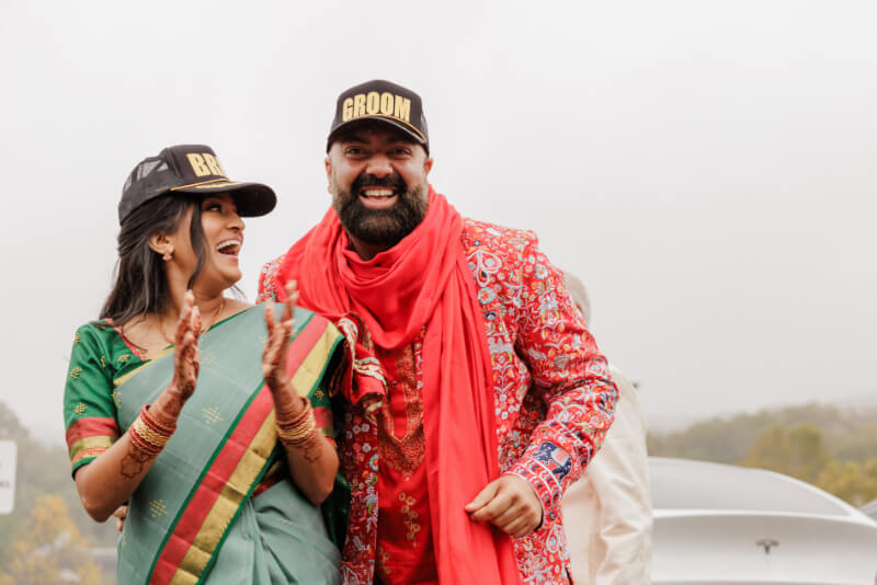 A woman in a green sari and a man in a red sherwani, both wearing hats labeled "Bride" and "Groom," stand together and smile outdoors.