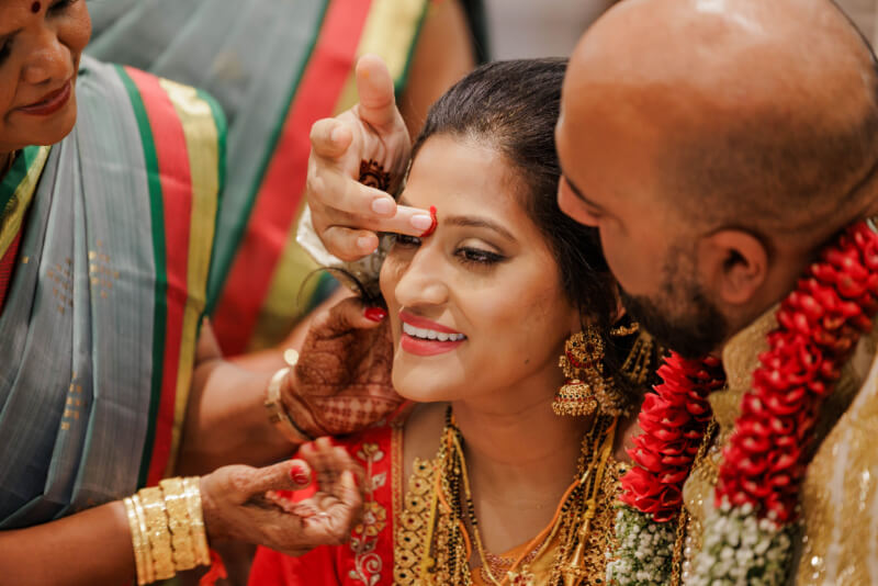 A woman in traditional attire smiles as another person applies a red mark to her forehead, with a man and another woman beside her during an Indian wedding ceremony.