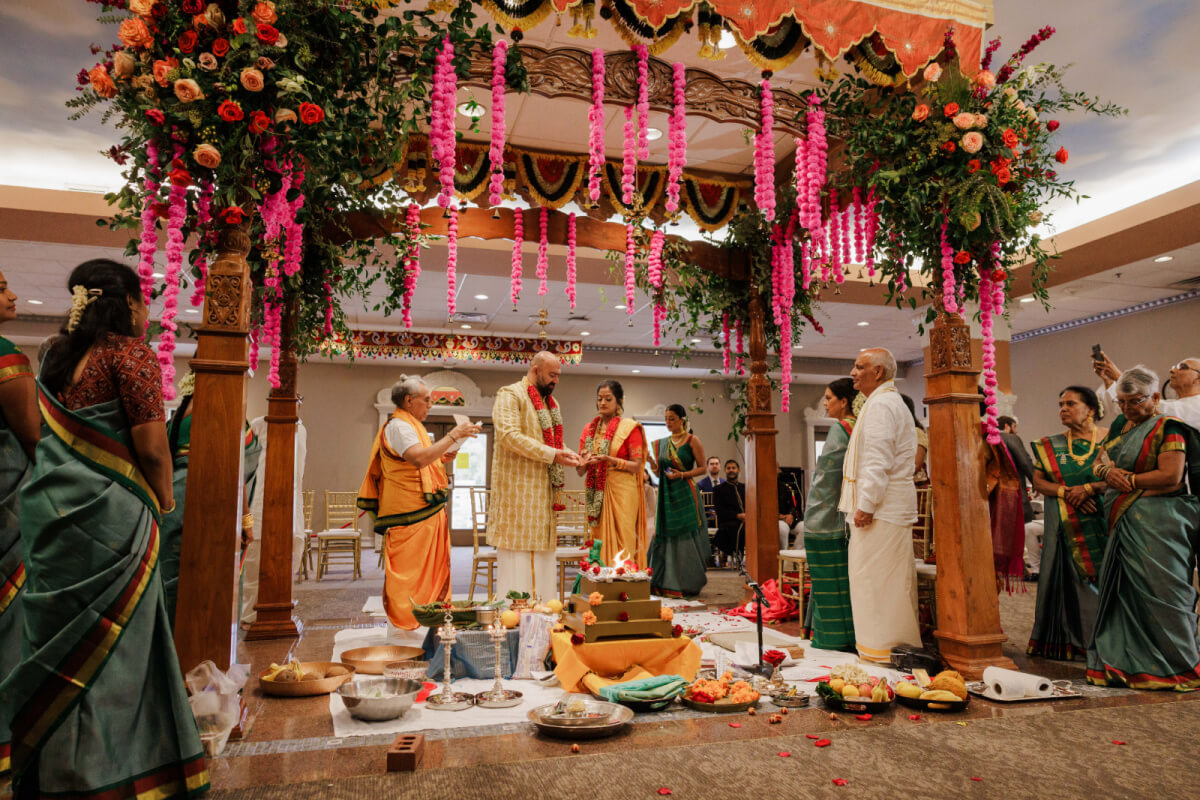 A couple stands under a decorated mandap during an Indian wedding ceremony, surrounded by family members in colorful attire and ritual items on the floor.
