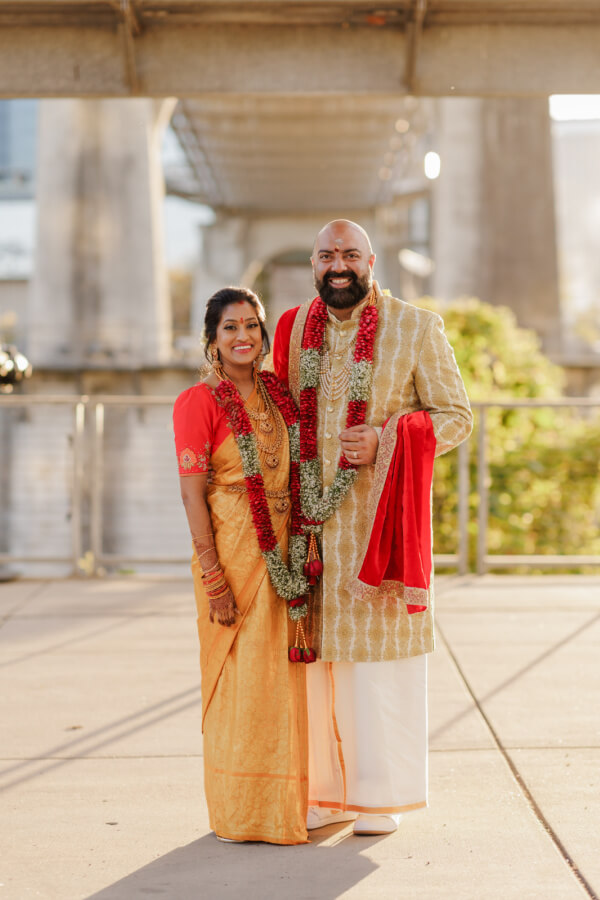 A man and woman in traditional Indian wedding attire, both wearing garlands, stand side by side outdoors under a bridge.