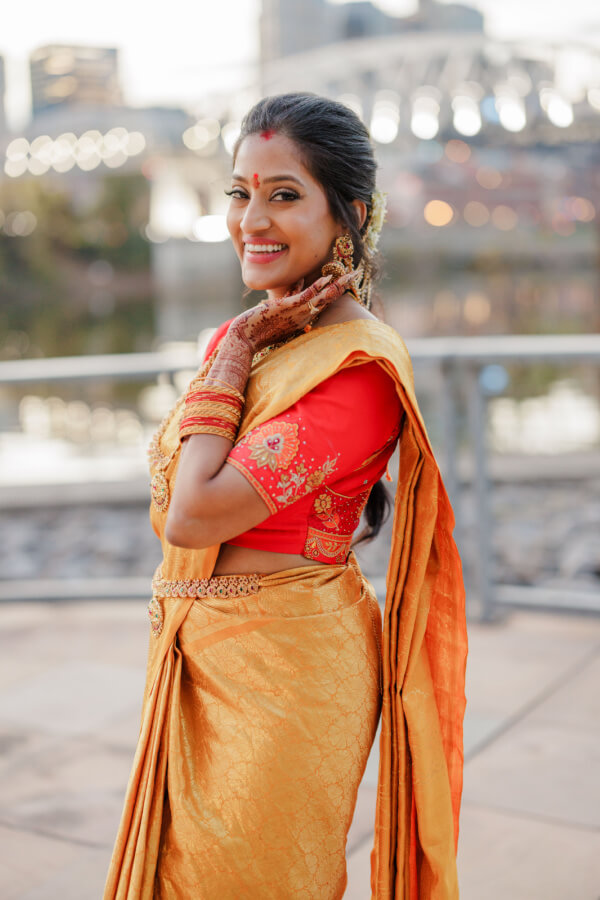 A woman in a traditional gold and red saree stands outdoors, smiling and touching her face, with a blurred cityscape in the background, capturing the joy of an Indian wedding.