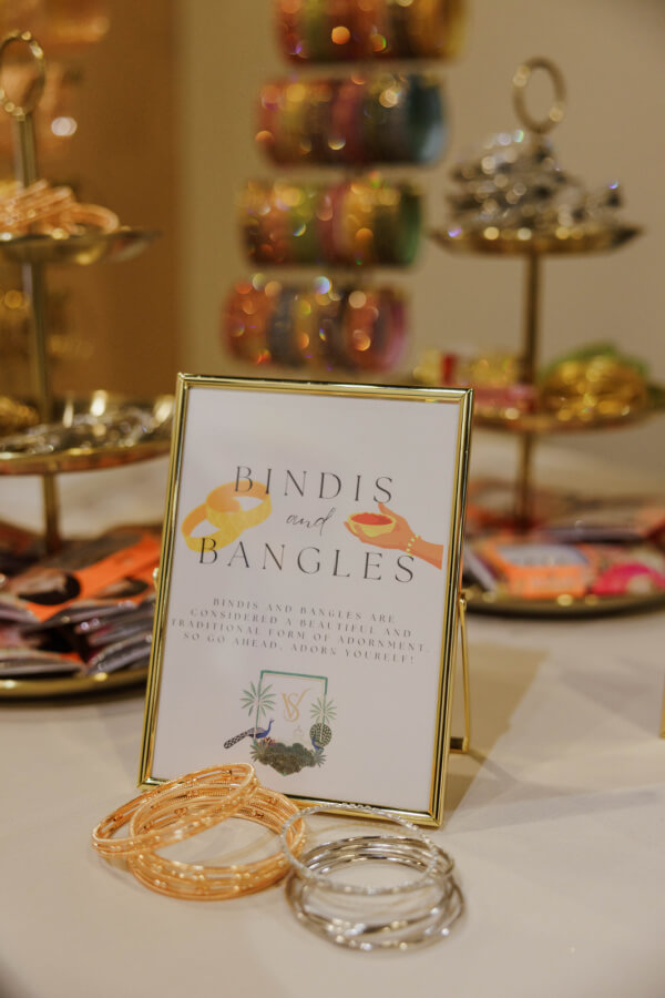 A table display with a sign reading “Bindis and Bangles,” featuring gold and silver glass bangles and colorful stacked bangle trays in the background.