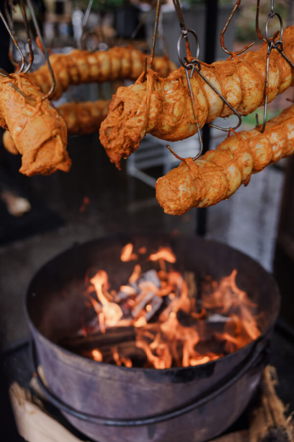 Several skewered sausages coated in a reddish seasoning are suspended over an open fire pit for roasting, reminiscent of the vibrant flavors often enjoyed at an Indian wedding.