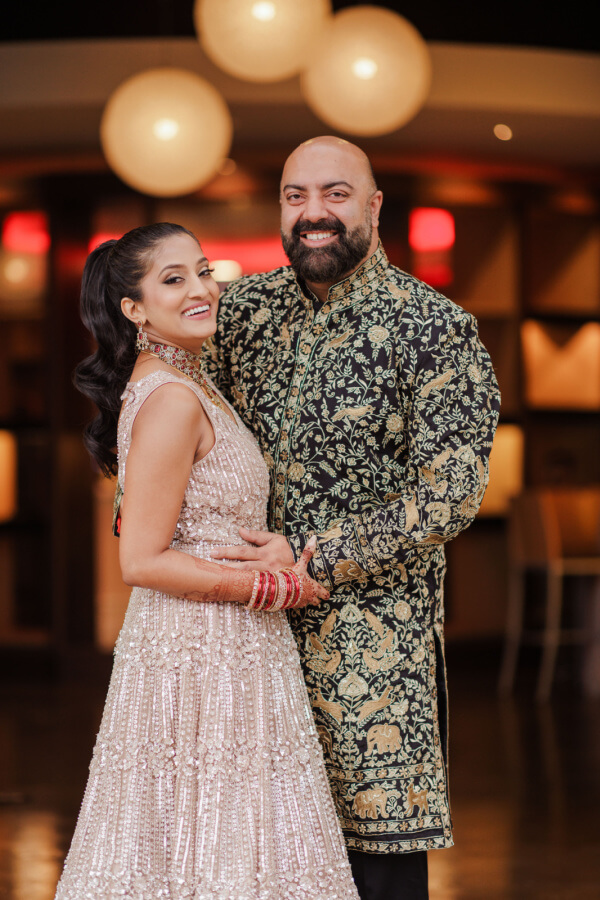 A smiling couple poses indoors at an Indian wedding, the woman in a light pink embroidered dress with red bangles, and the man in a black and gold patterned sherwani.