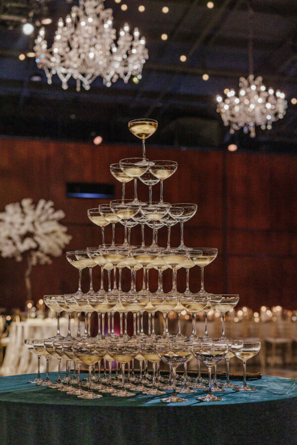 A pyramid of stacked champagne glasses filled with champagne, set on a table under crystal chandeliers at an elegant Indian wedding event venue.