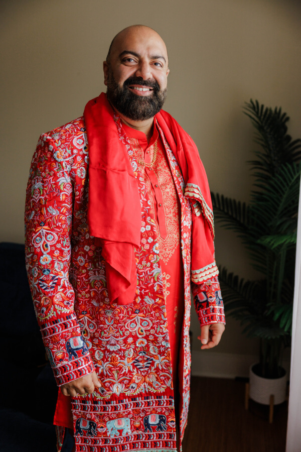 A man with a trimmed beard smiles while wearing an ornate red traditional outfit with intricate patterns, standing indoors next to a potted plant, ready for his wedding.