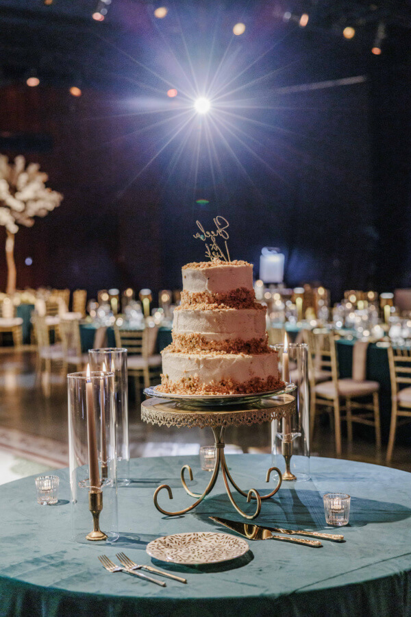A three-tiered cake with gold accents sits on a decorative stand on a round table, surrounded by candles in a dimly lit banquet hall, capturing the elegance of an Indian wedding celebration with beautifully arranged tables and chairs.