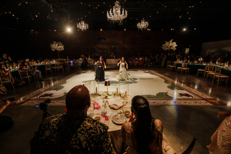 Two women perform in traditional attire on a large decorated dance floor at an Indian wedding, observed by seated guests in a dimly lit, elegant hall with chandeliers.