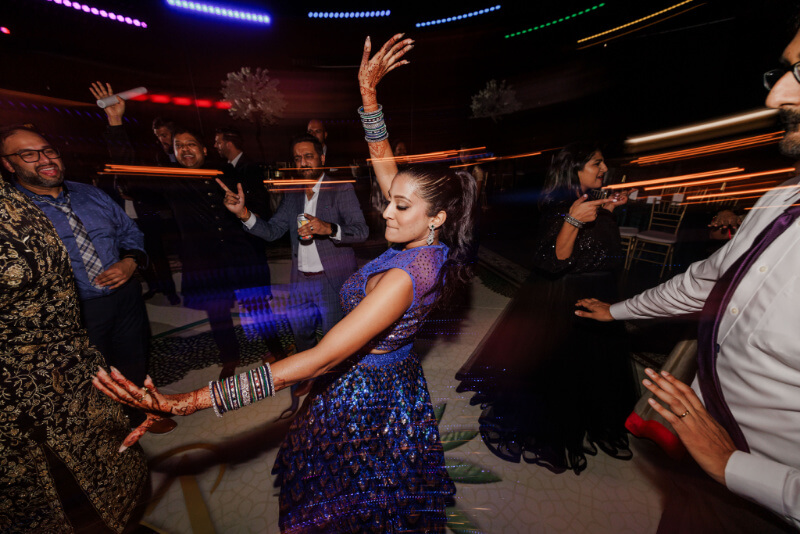 A woman in a blue dress dances energetically at a lively indoor Indian wedding party, surrounded by other guests, colorful lights, and motion blur in the background.