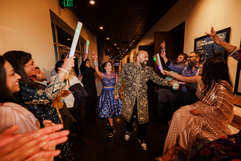 A couple in festive attire walks down a hallway at an Indian wedding, smiling and holding hands, while a group of people around them cheer and raise glow sticks.
