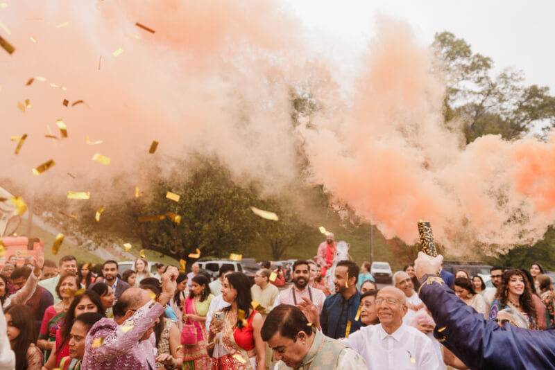 A crowd of people in colorful attire celebrates outdoors at an Indian wedding, with orange smoke and gold confetti filling the air.