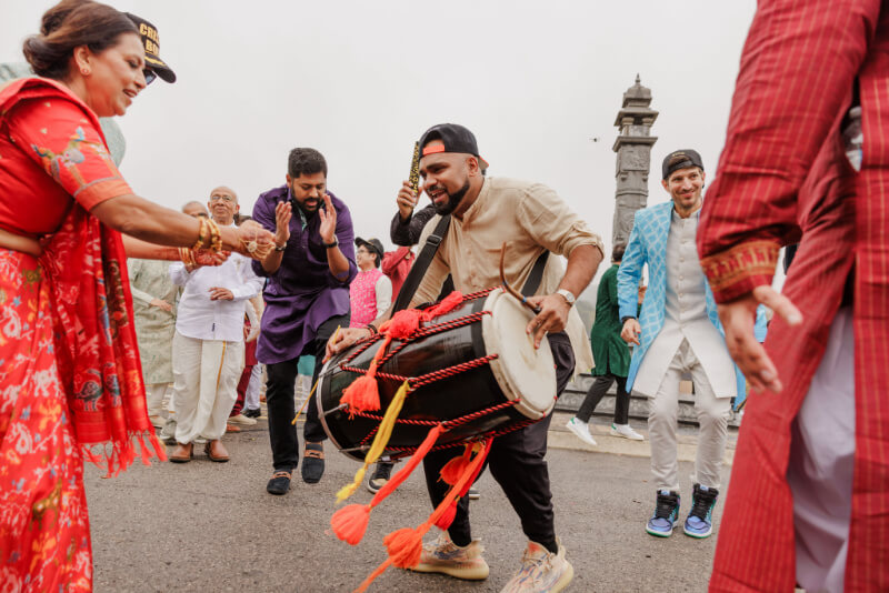A group of people in colorful traditional attire dance and play a dhol drum outdoors during a festive Indian wedding celebration.