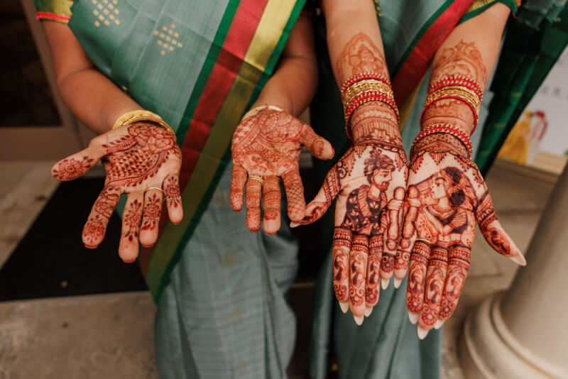 Two people in green sarees display intricate mehndi designs on their palms, featuring detailed patterns and portraits, with gold bangles adorning their wrists—capturing the vibrant spirit of an Indian wedding.