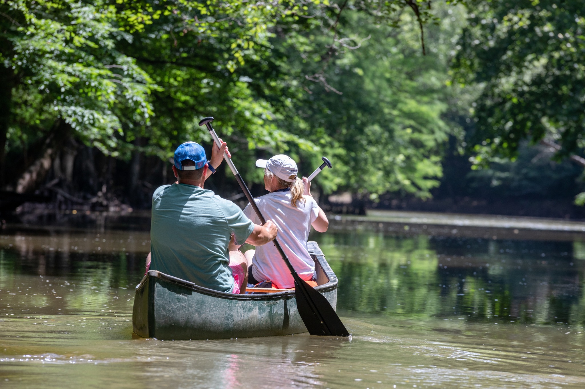Two people paddle a canoe down a calm, tree-lined river on a sunny day, exploring the scenic beauty often found near Mississippi lakes.