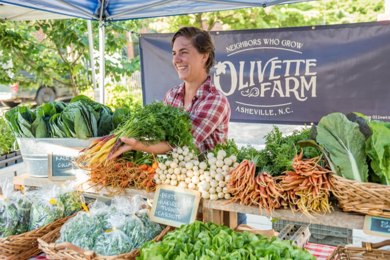 A vendor smiles behind a table of fresh vegetables, including carrots, beets, and greens, at an outdoor Olivette Farm stand.